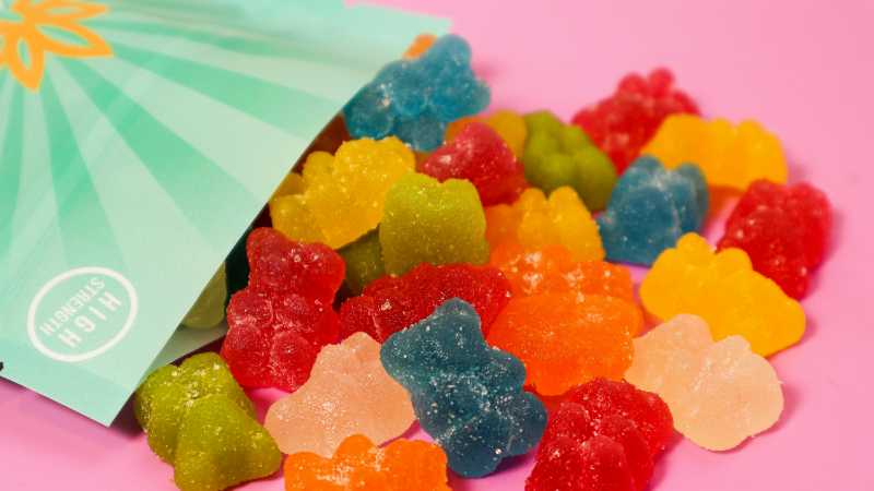 Close-up of colorful gummy bears spilling out of a high-strength package on a pink surface., tags: ein zum - pexel